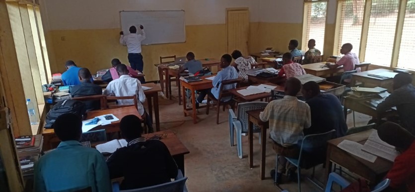 People sat at desks in a classroom while a teacher writes on the white board at the front