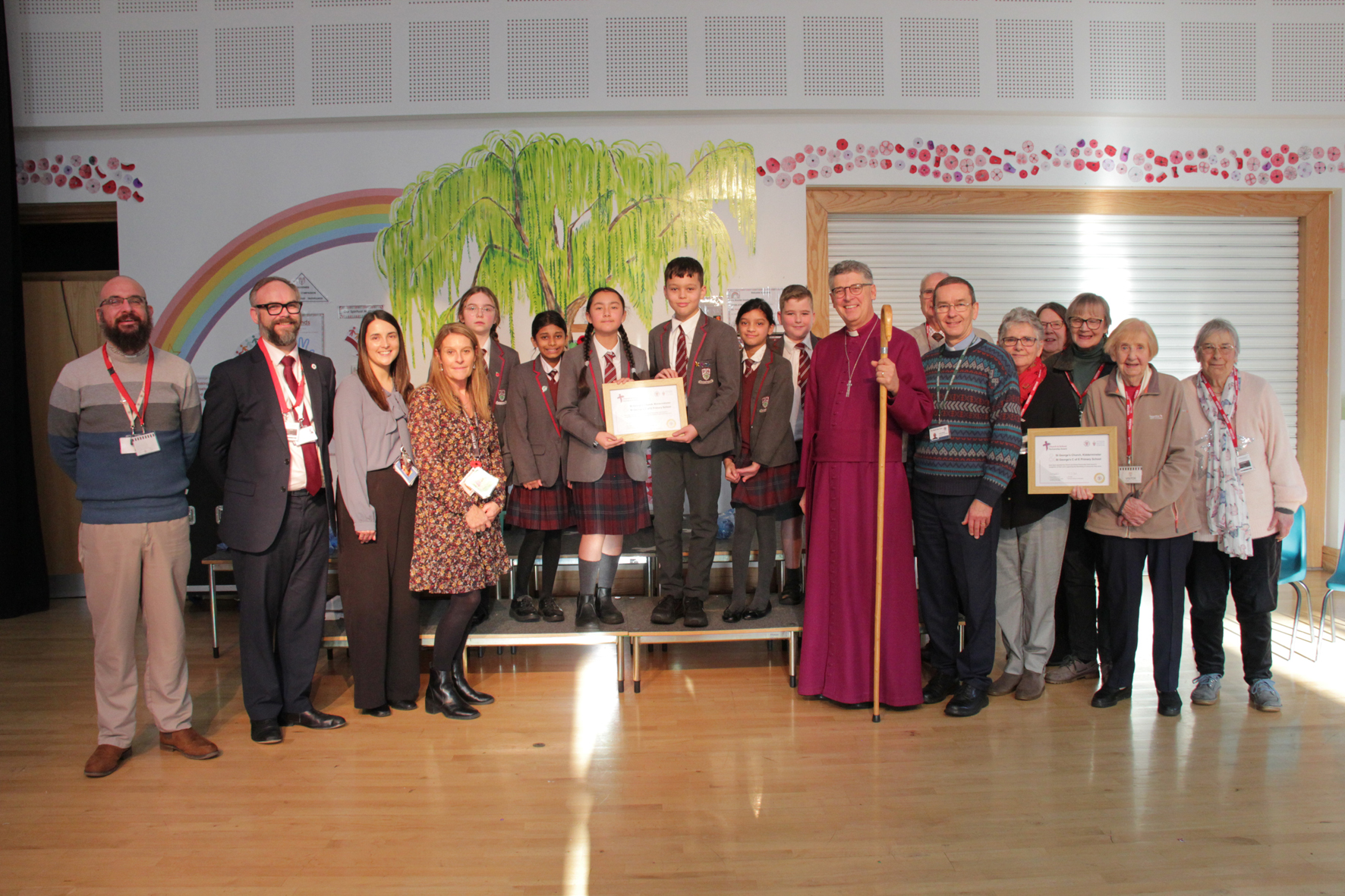 A group of adults and children standing in the school hall holding two award certificates