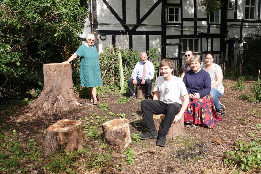 Team members sitting on logs in the outdoor worship area as Sue Adeney stands next to the tree stump altar