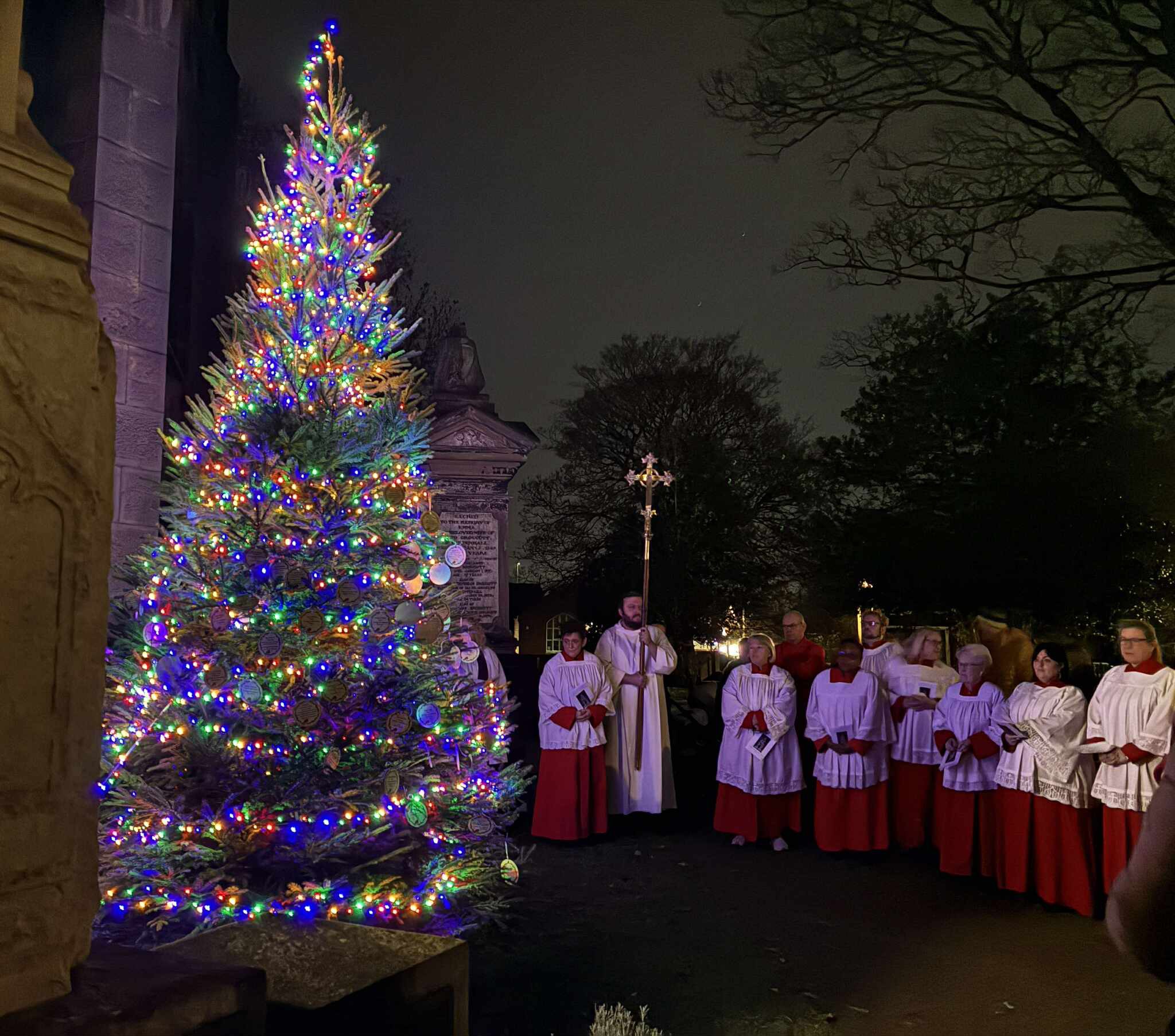 Choir stood outside the church next to a big Christmas tree lit up with multi-coloured lights