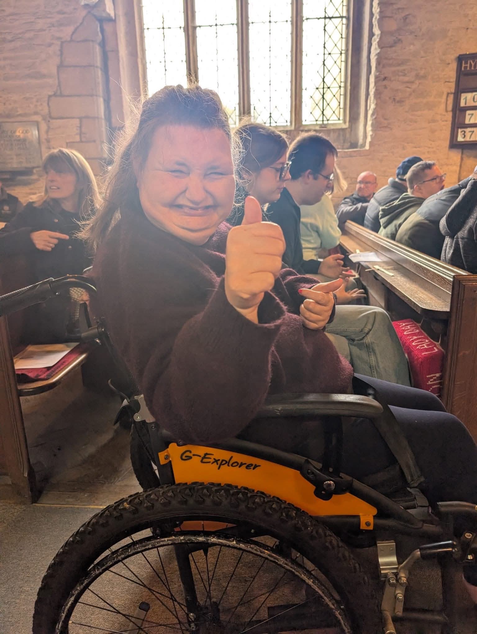 A lady giving a thumbs up and sitting in a wheelchair next to a row of people in a pew