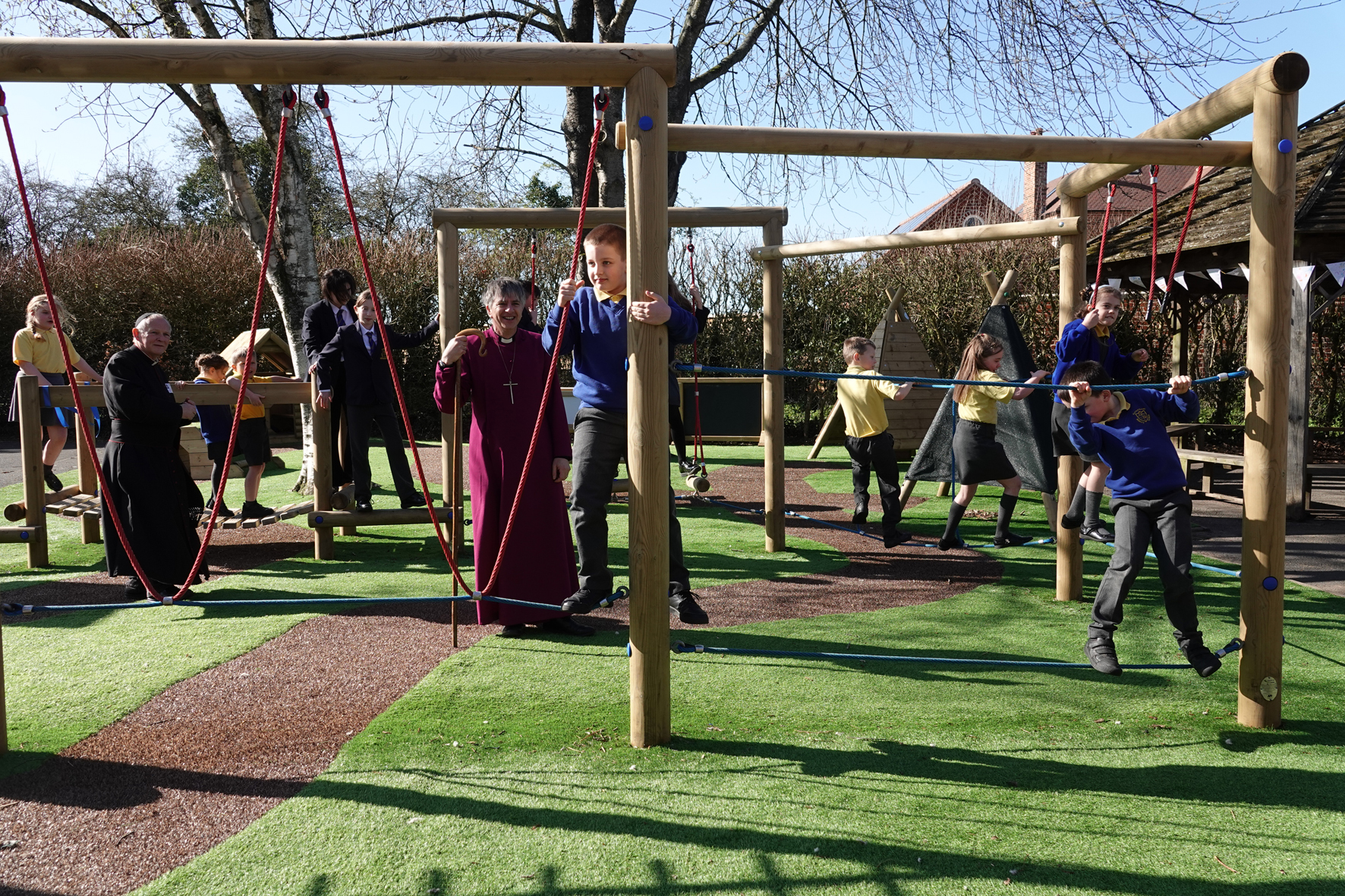 Bishop Hugh standing in the middle of a set of wooden play equipment while children climb around him.