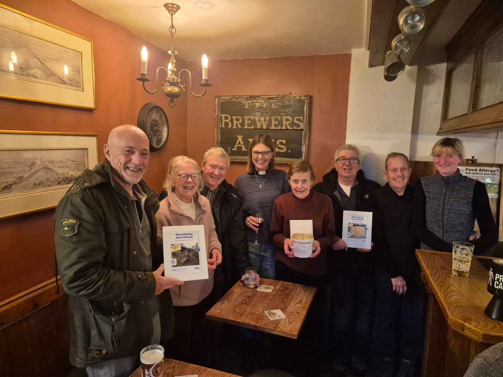 A group of people standing in a pub holding three toilet twinning certificates