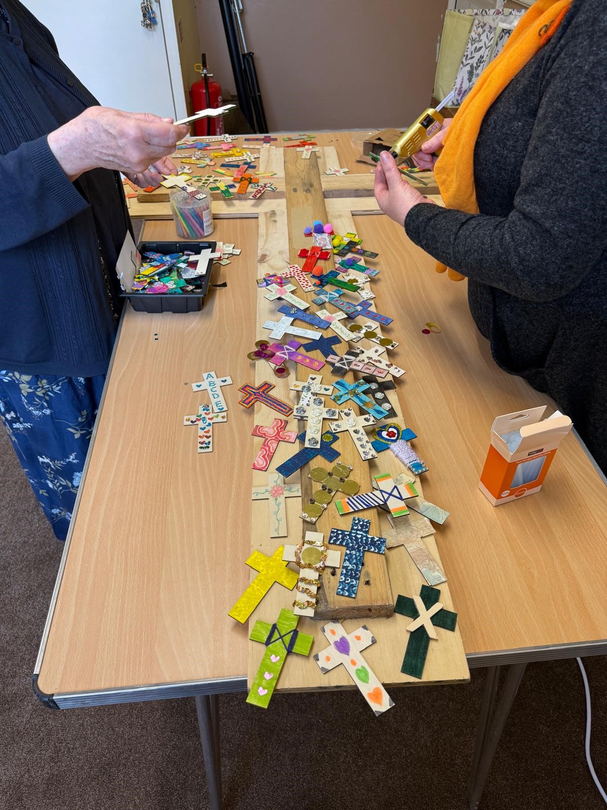A wooden cross on the table as two people stick smaller decorated crosses on it.