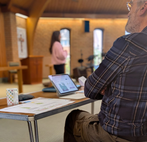 Person sitting in a church listening to a lady speaking at the front with his ipad with stats on the screen