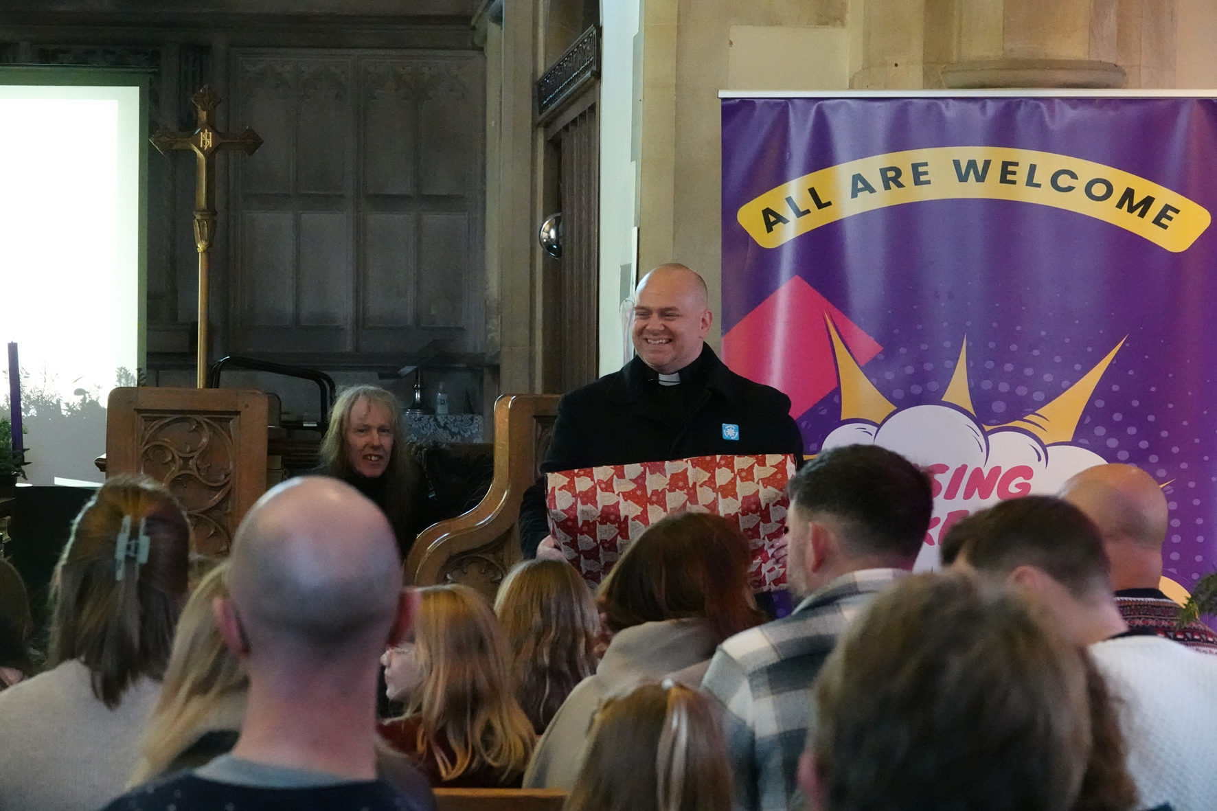 Shaun Armstrong stands in front of a congregation holding a big parcel wrapped in Christmas paper