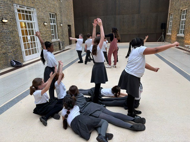 Children from St Barnabas school sitting, laying and standing in a hall as they take part in a drama workshop