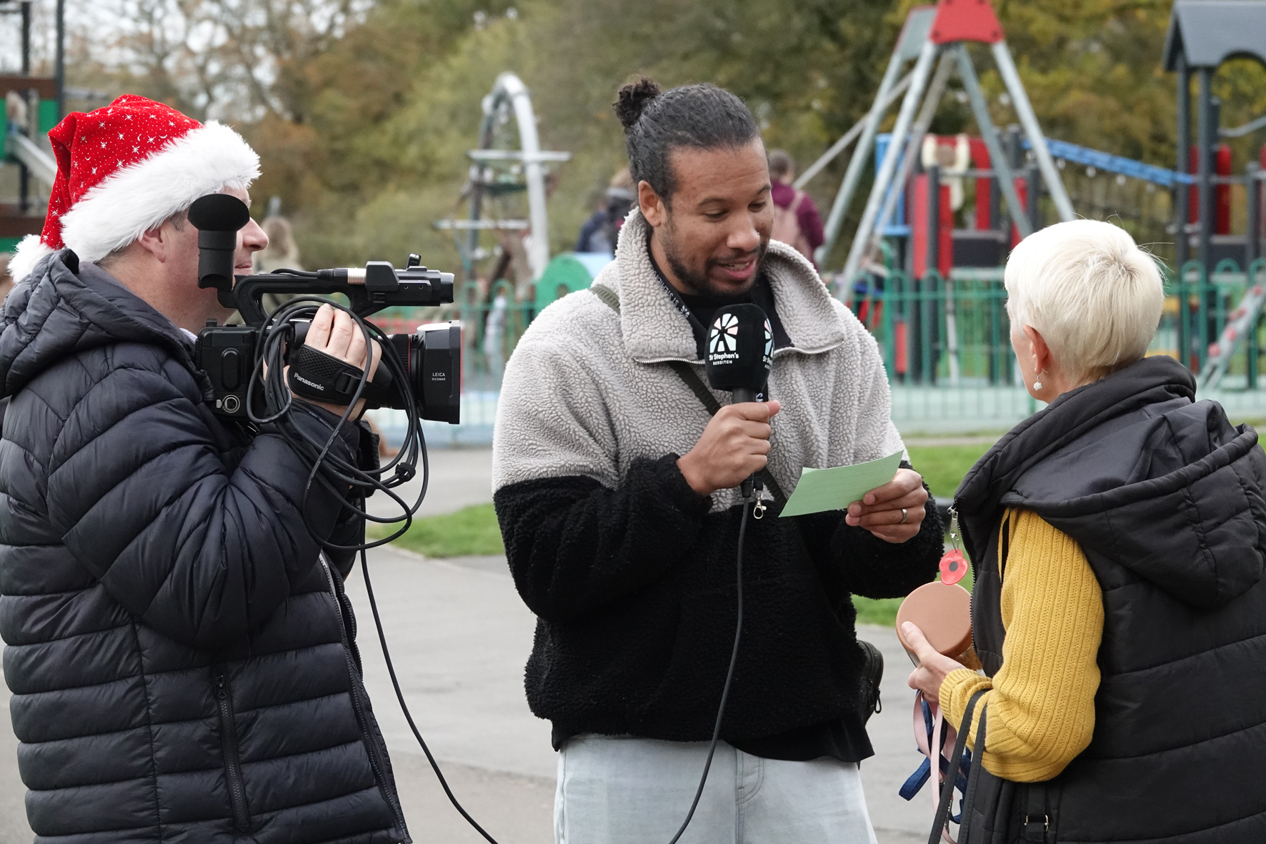 Person in a santa hat holding a camera with another person holding a microphone interviewing a lady