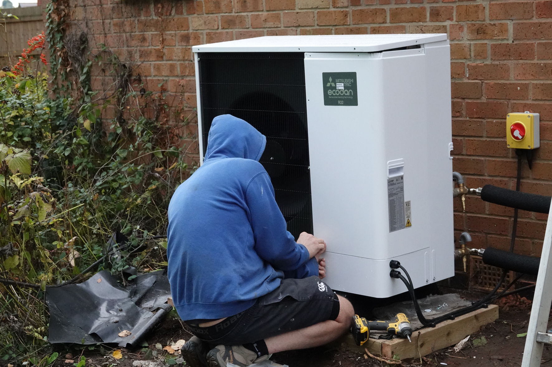 Workman kneeling in front of a heat pump as he installs it on the side of a house