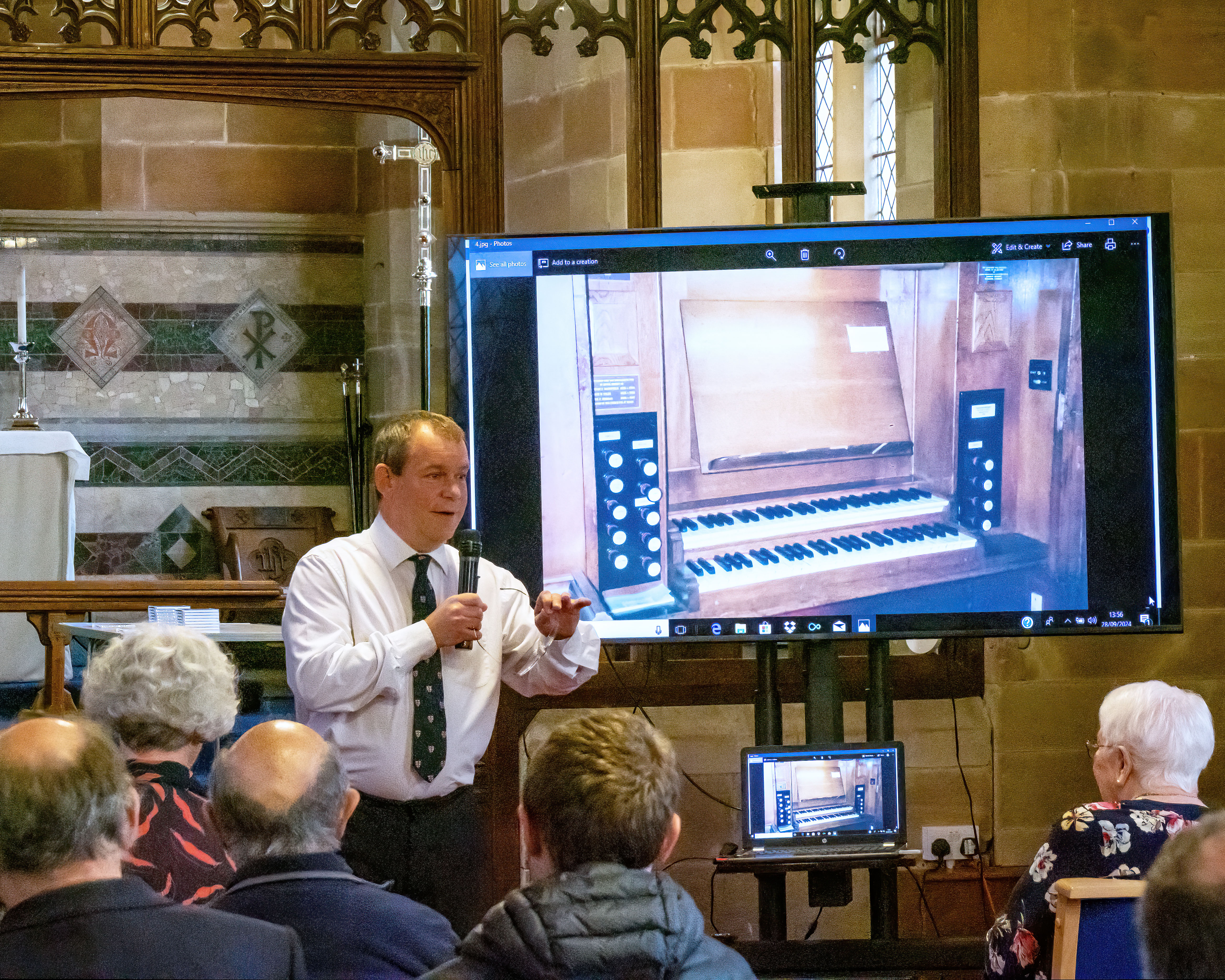 Man stood at the front of webheath church with a screen showing the organ and talking into a microphone