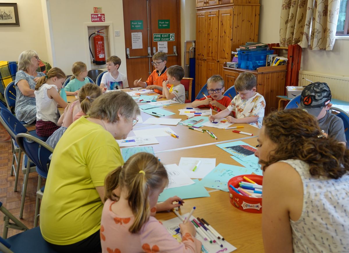 The children supported by three adults sit round one large table colouring in a handout.