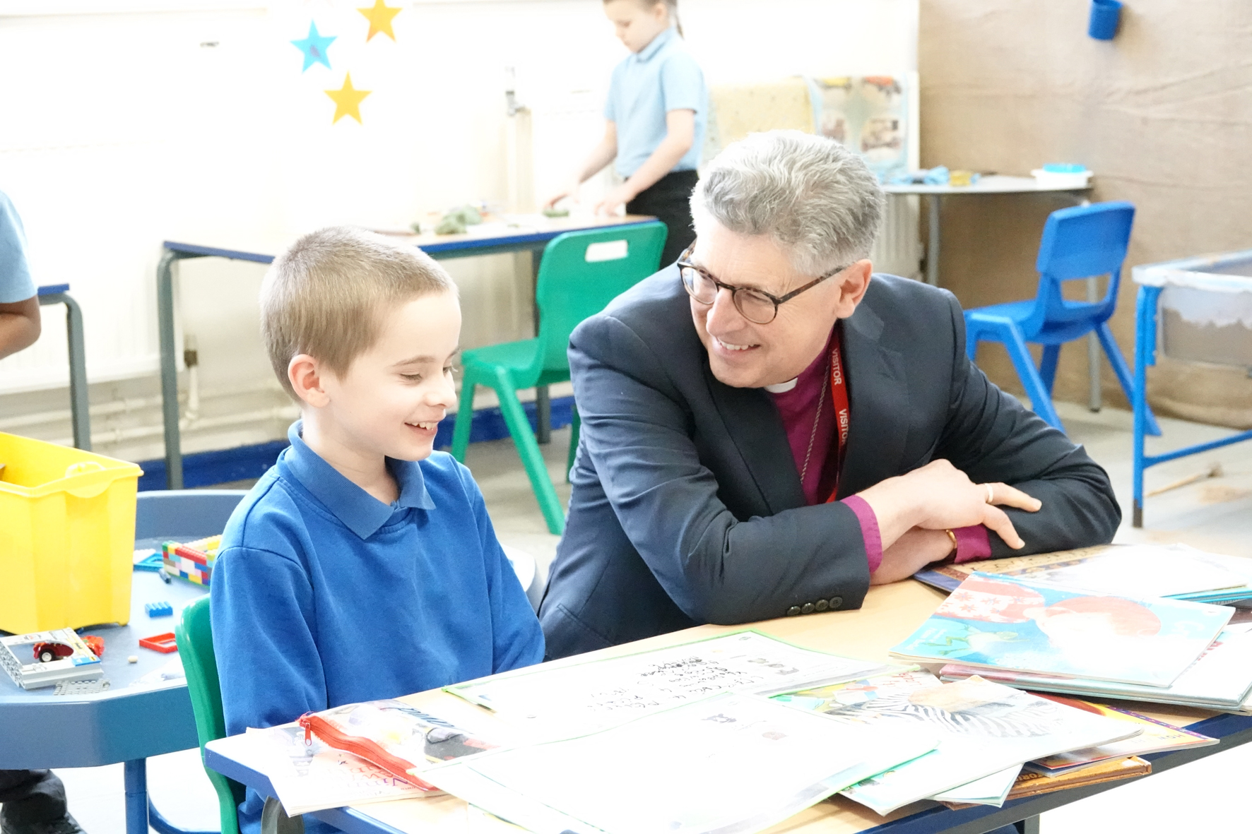 Bishop Martin crouches next to a pupil who is sitting at a desk, looking together at the work they are doing