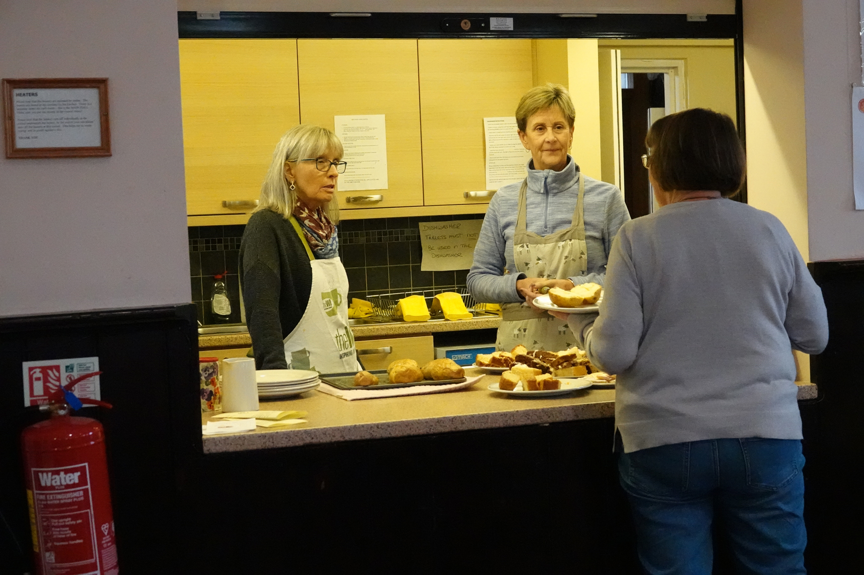 Two people standing behind a serving hatch in a kitchen with cakes on the counter as they serve the person in front