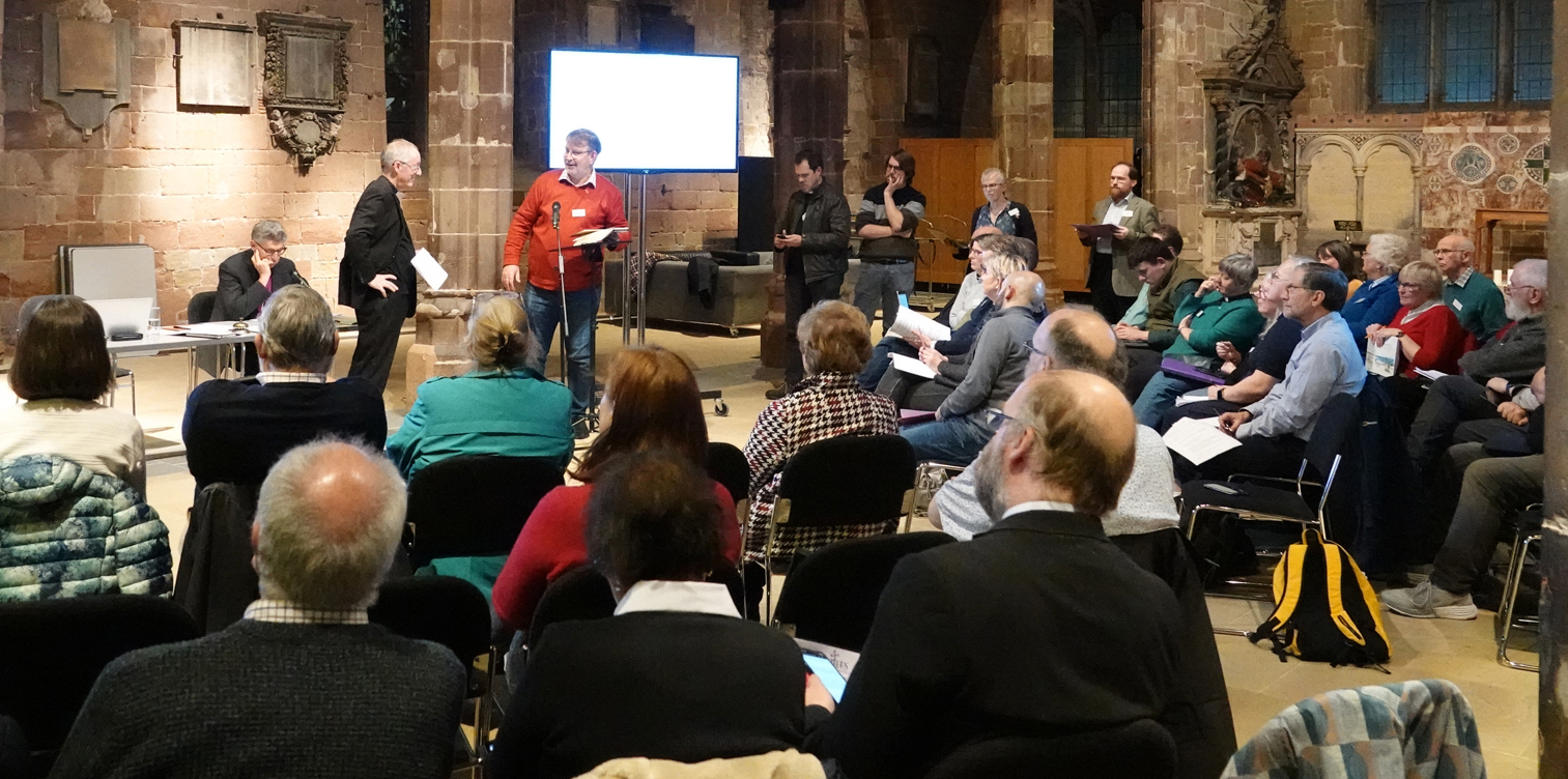 People standing up to speak at diocesan synod with others sat in a semi circle listening