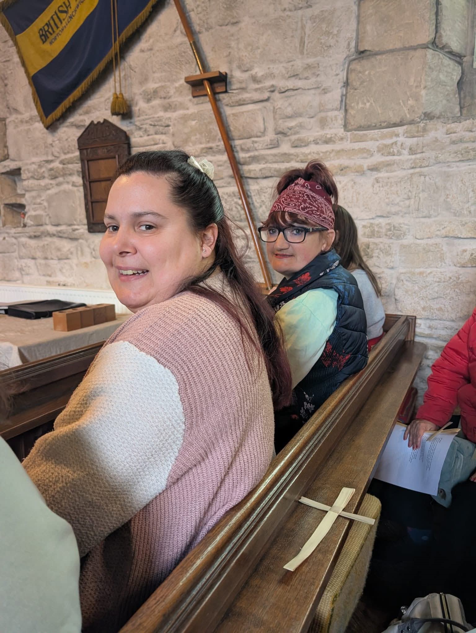 Two ladies looking over their shoulder as they sit in a pew with a palm cross on the pew rest