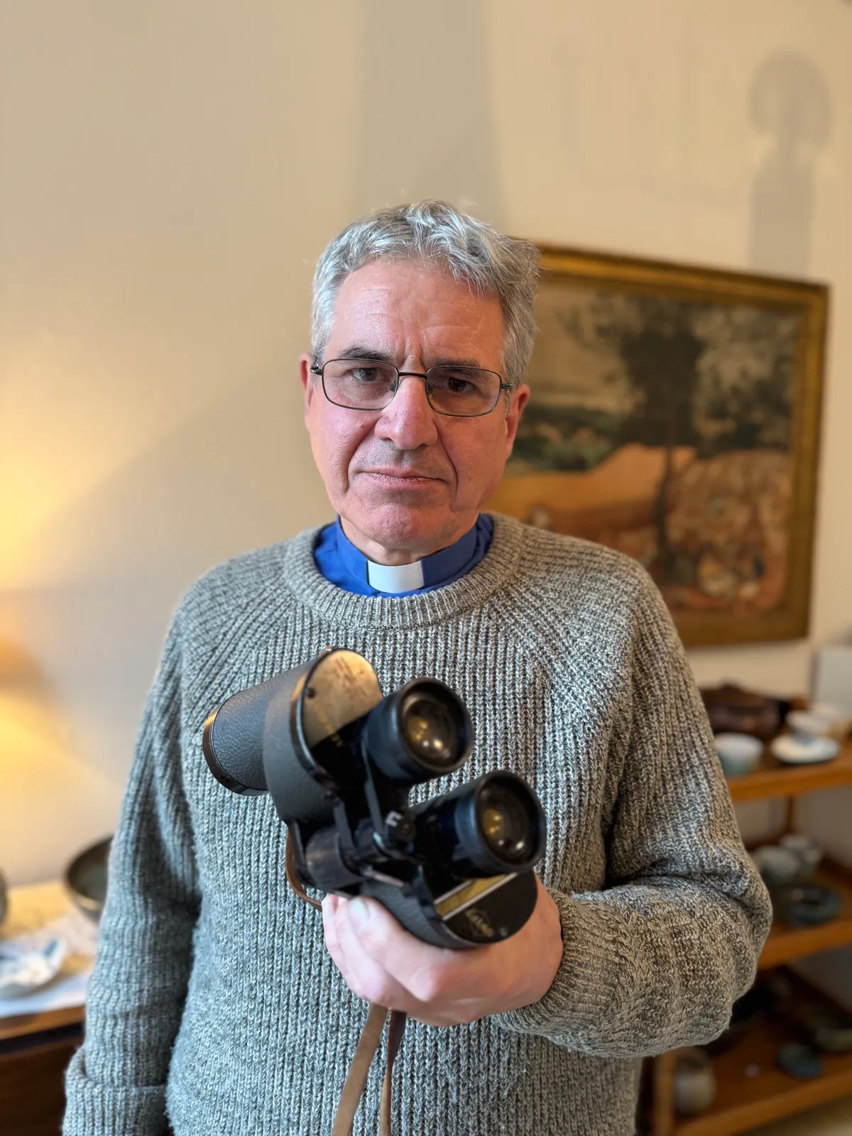 John Flitcroft standing in his home holding a pair of binoculars