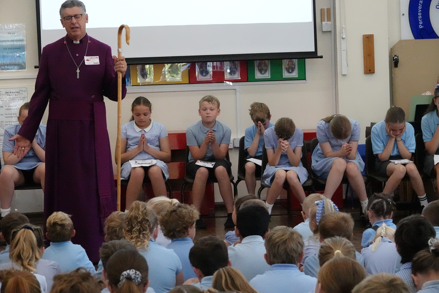 Bishop Martin in his cassock and shepherds crook saying a prayer in an assembly with the children praying
