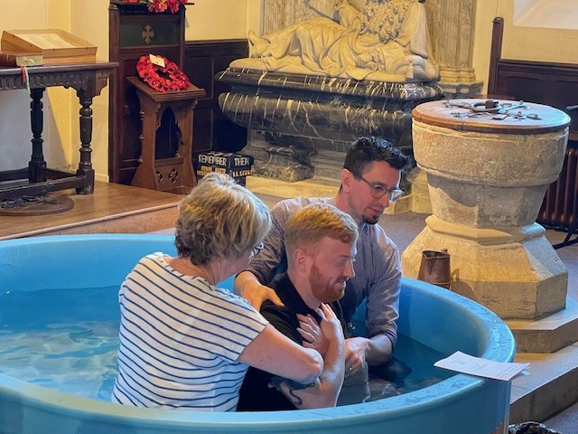 Man kneeling in a font with his arms crossed over with a person either side of him about to conduct a full emersion baptism