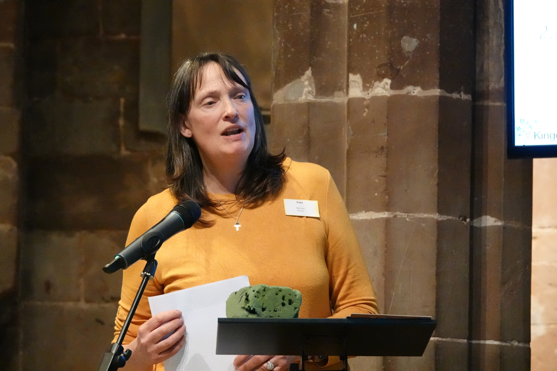 Kate Hurst standing at the microphone at diocesan synod with a piece of floral foam on the lecturn