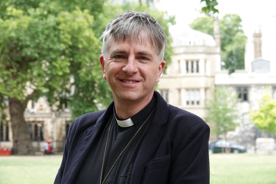 Head and shoulders shot of Bishop Hugh Nelson standing in front of a green space with a tree and building in the background