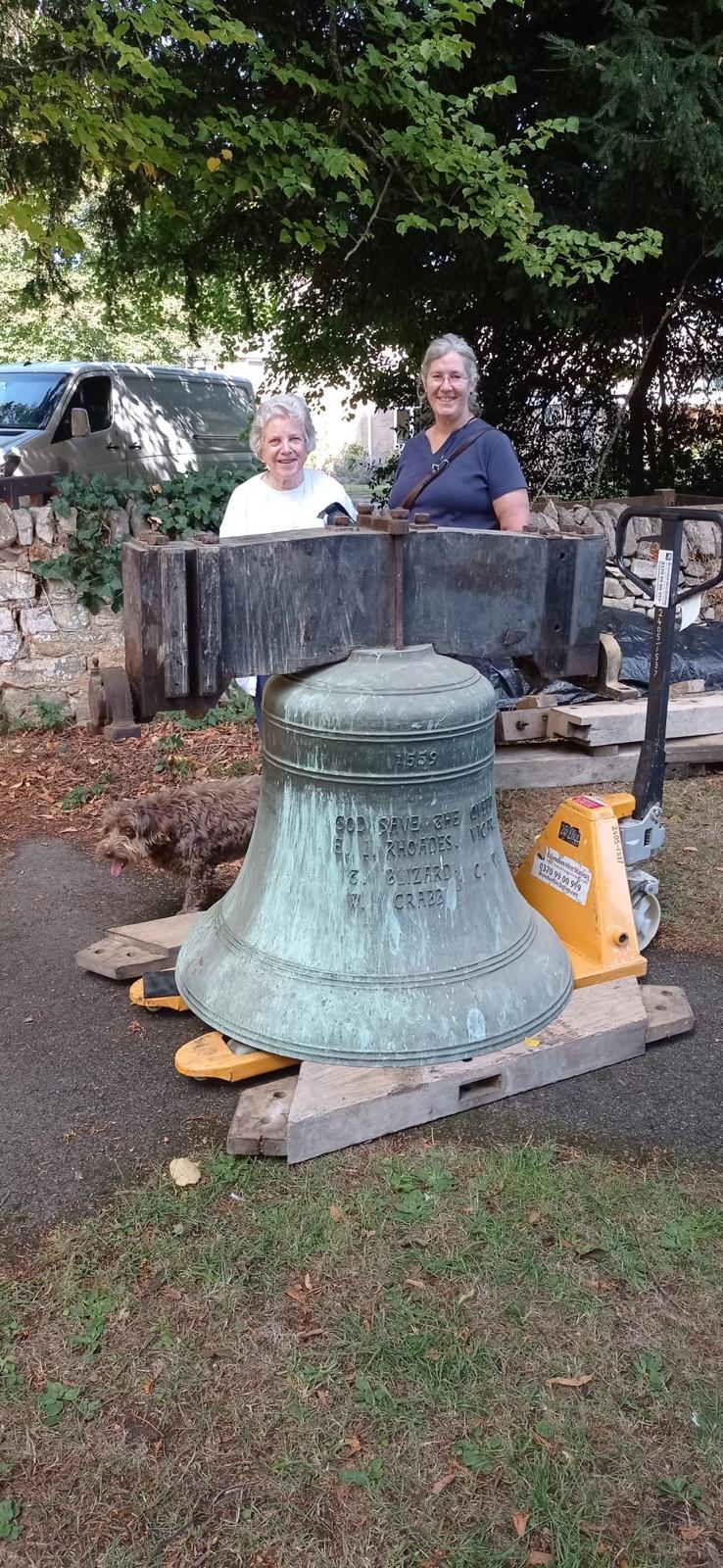 A bell resting on two blocks of wood on the floor with two ladies standing behind