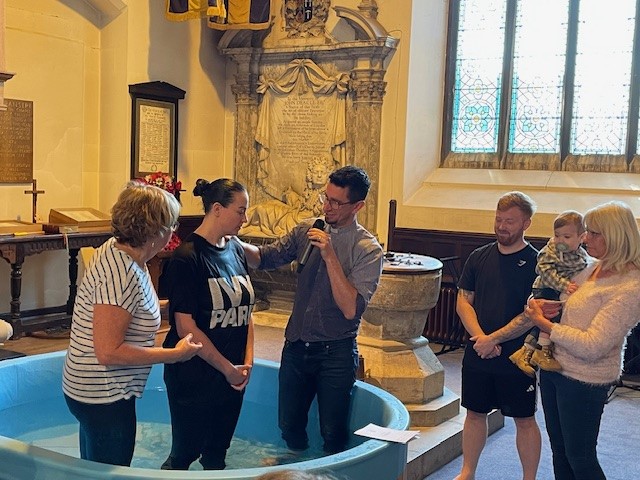 Lady stood in a large baptismal font flanked by 2 people, with one speaking into a microphone. Watching are the lady's husband and a lady holding the couple's child