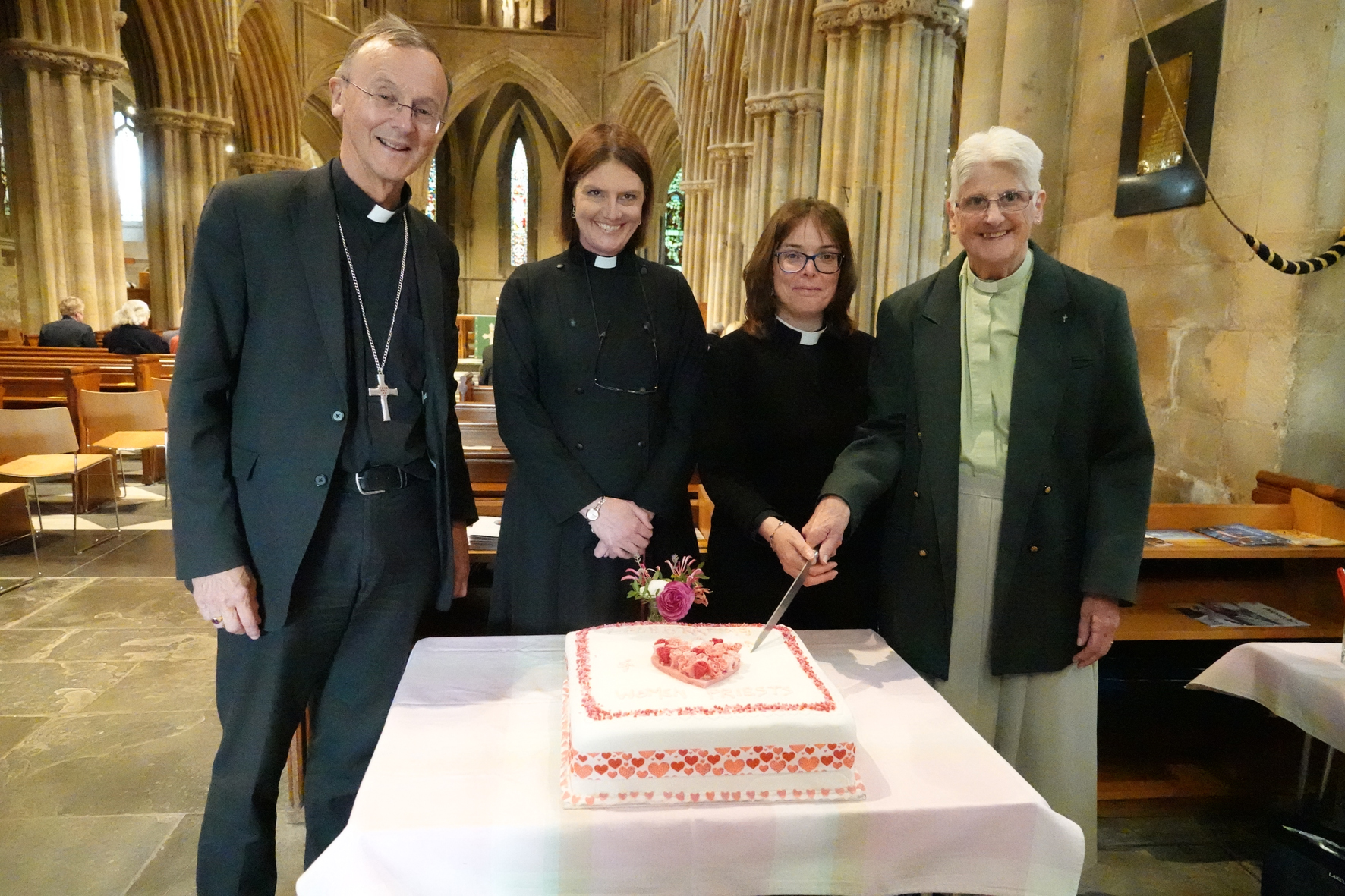 Bishop John standing with Becky Elliot as Jayne Parker and Andi Jones cut a big cake.