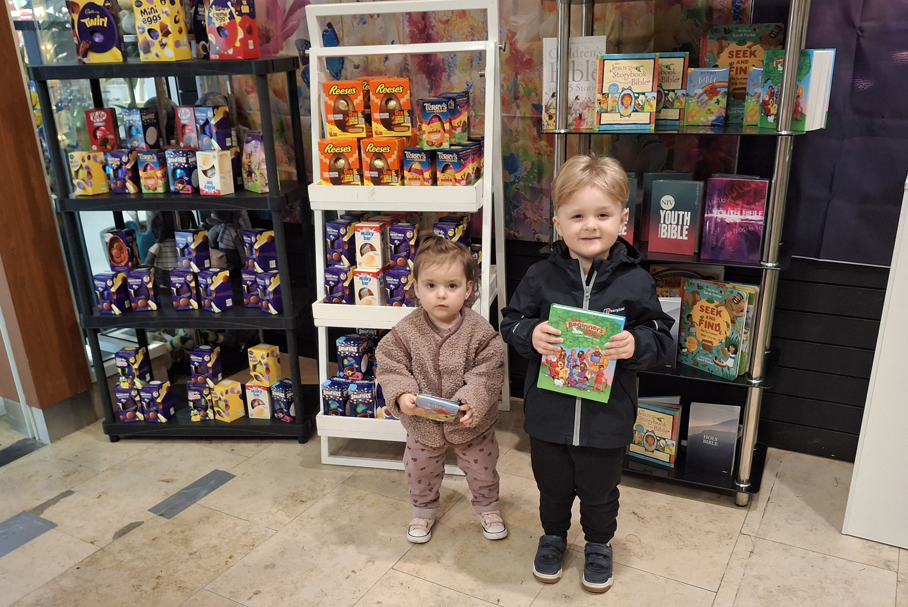 Two small children holding an Easter egg and bible standing in front of a display of the same