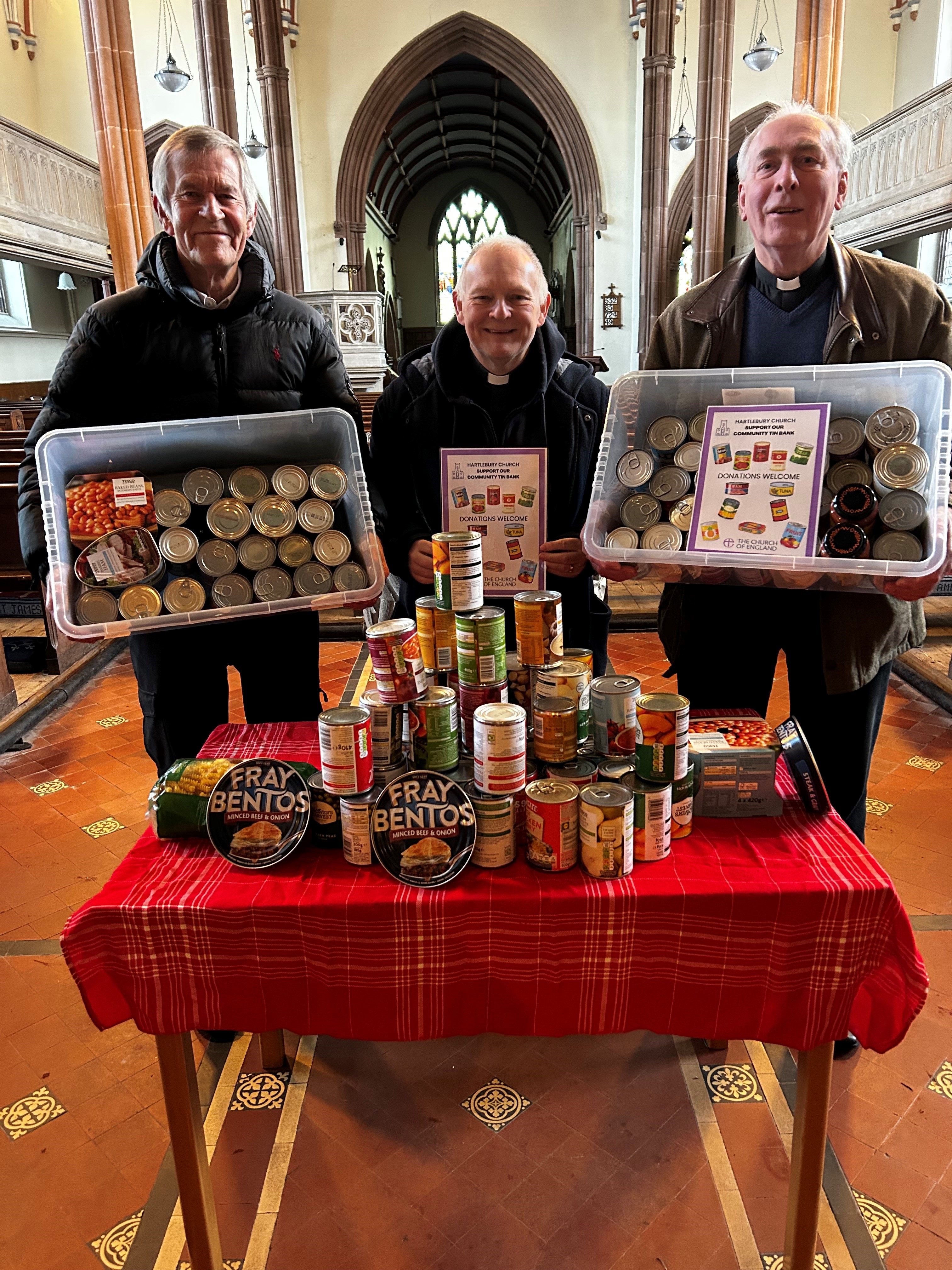 Three men holding boxes full of tins and standing behind a table of tins inside Hartlebury Church