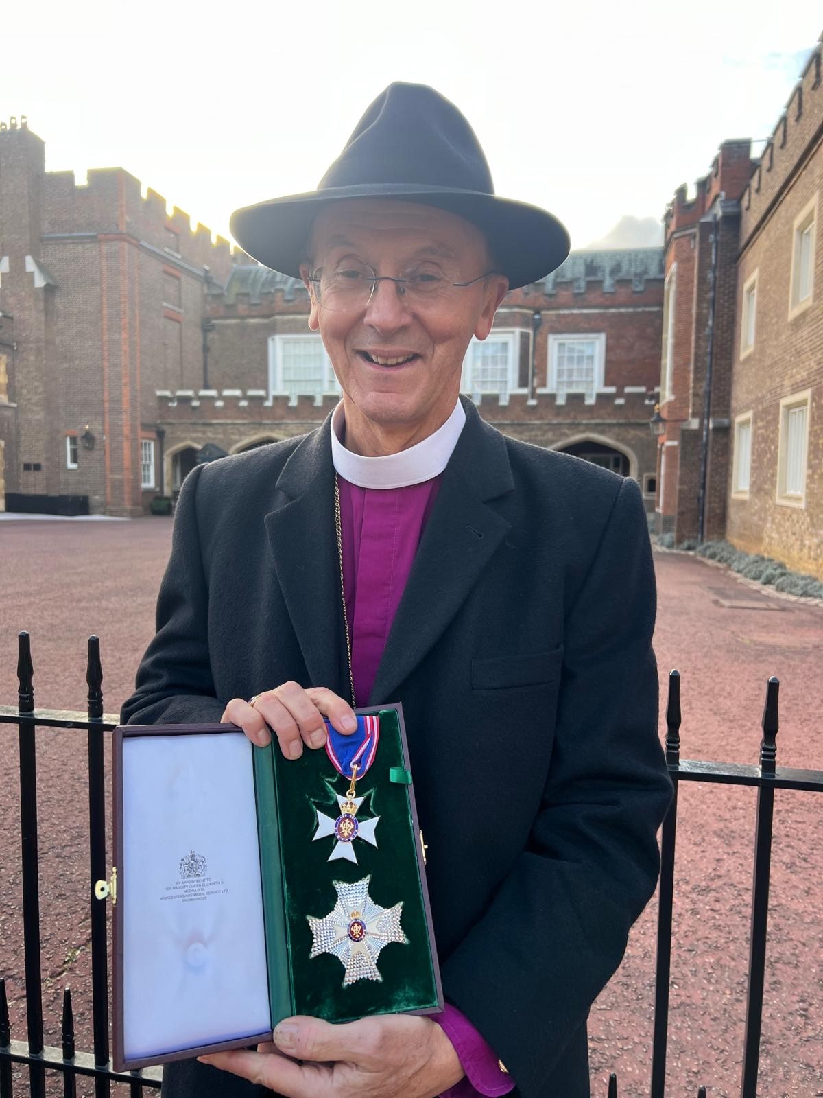 Bishop John standing holding his Knighthood from King Charles in a presentation box