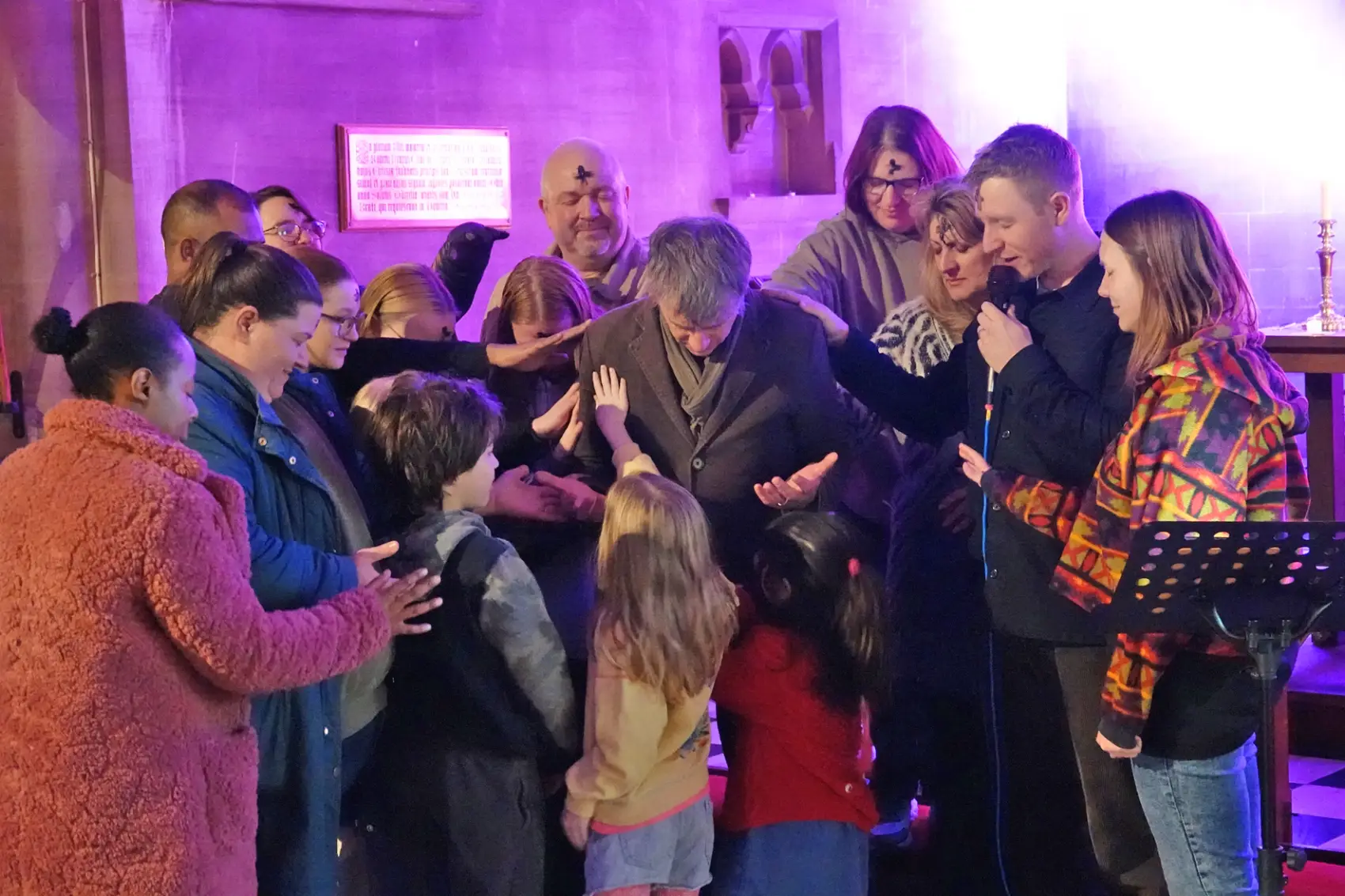 A group of people, both adults and children standing around Bishop Hugh as they pray for him.