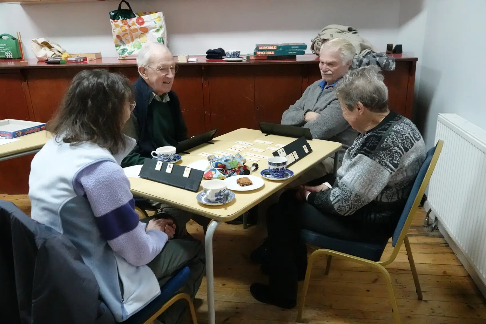 Four people sitting around a table playing Rummikub with tea and cake