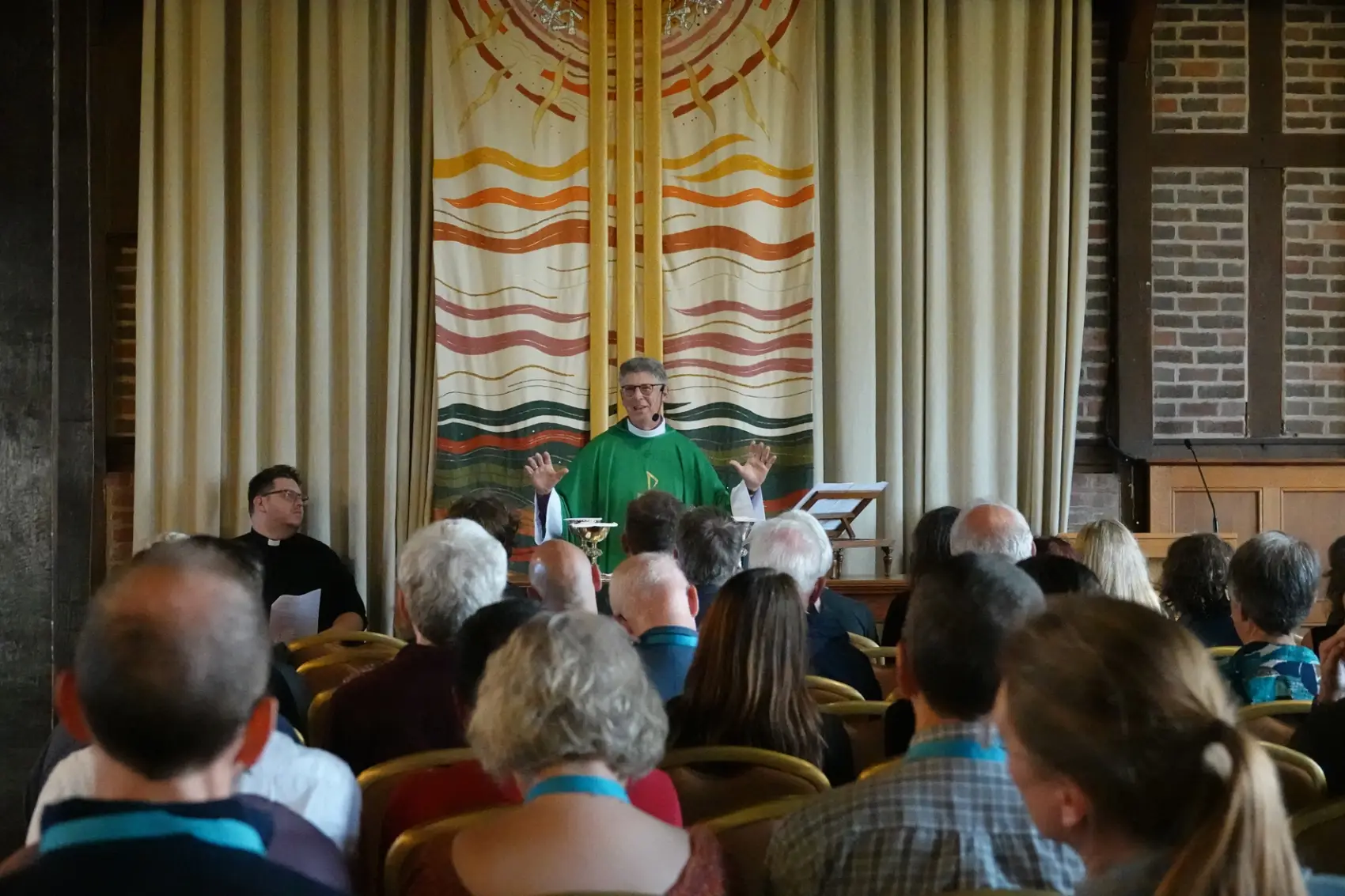Bishop Martin in his robes standing at the front of the chapel while people sit on chairs looking at him. 