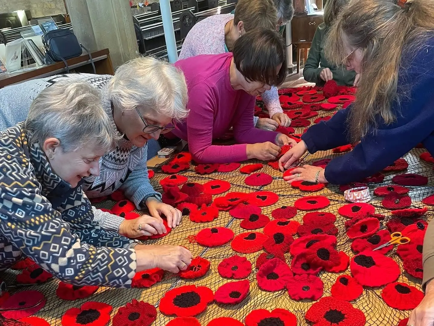 A group of ladies bending over a table of knitted poppies sewing them onto netting