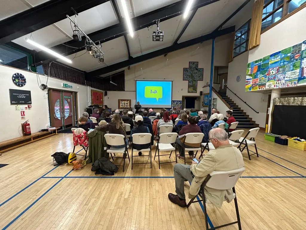 Photo taken from the back of a school hall where people sit in rows in front of a screen with 'talk' on it