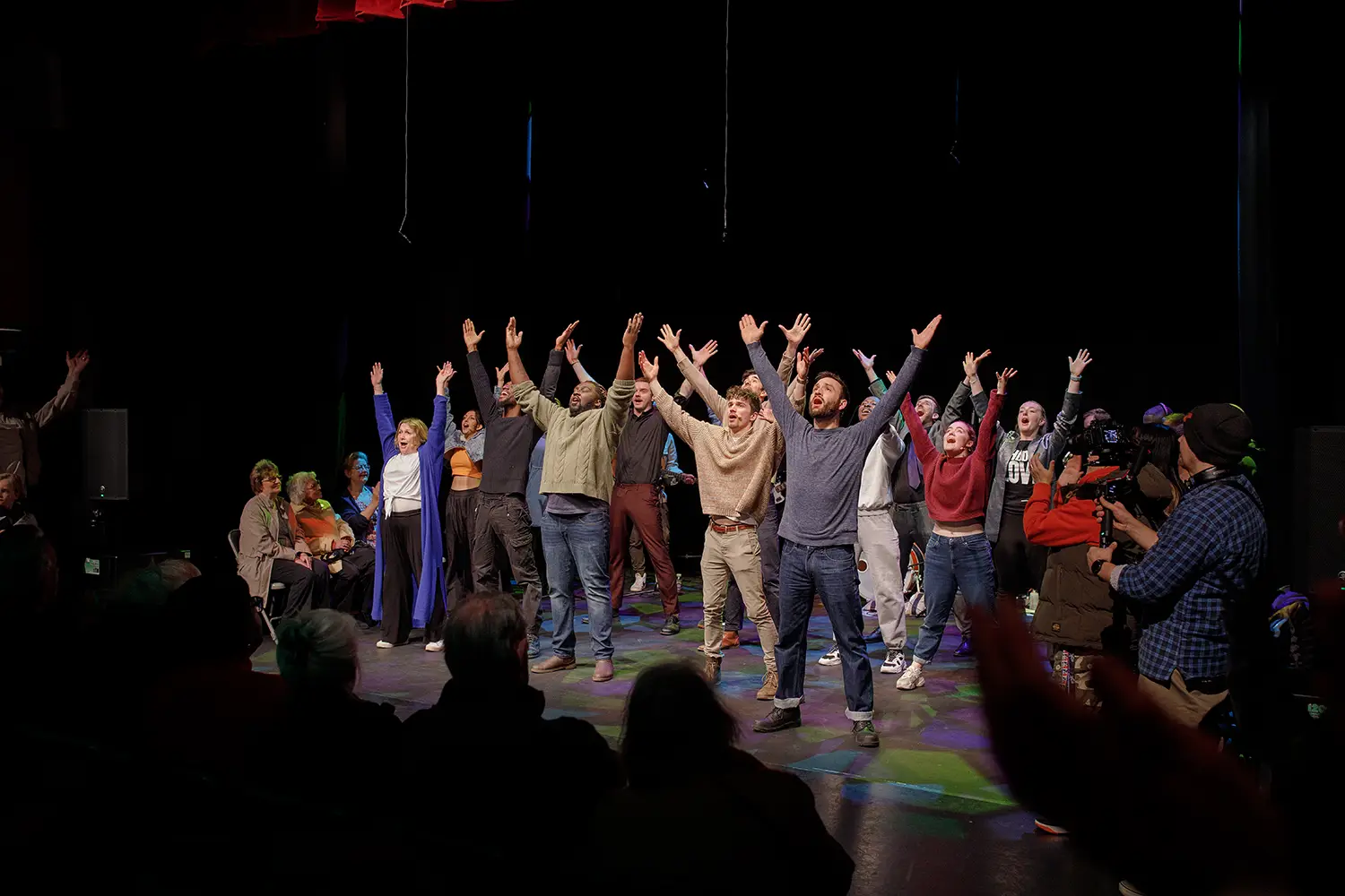 A group of actors standing on a stage in darkness with their hands in the air