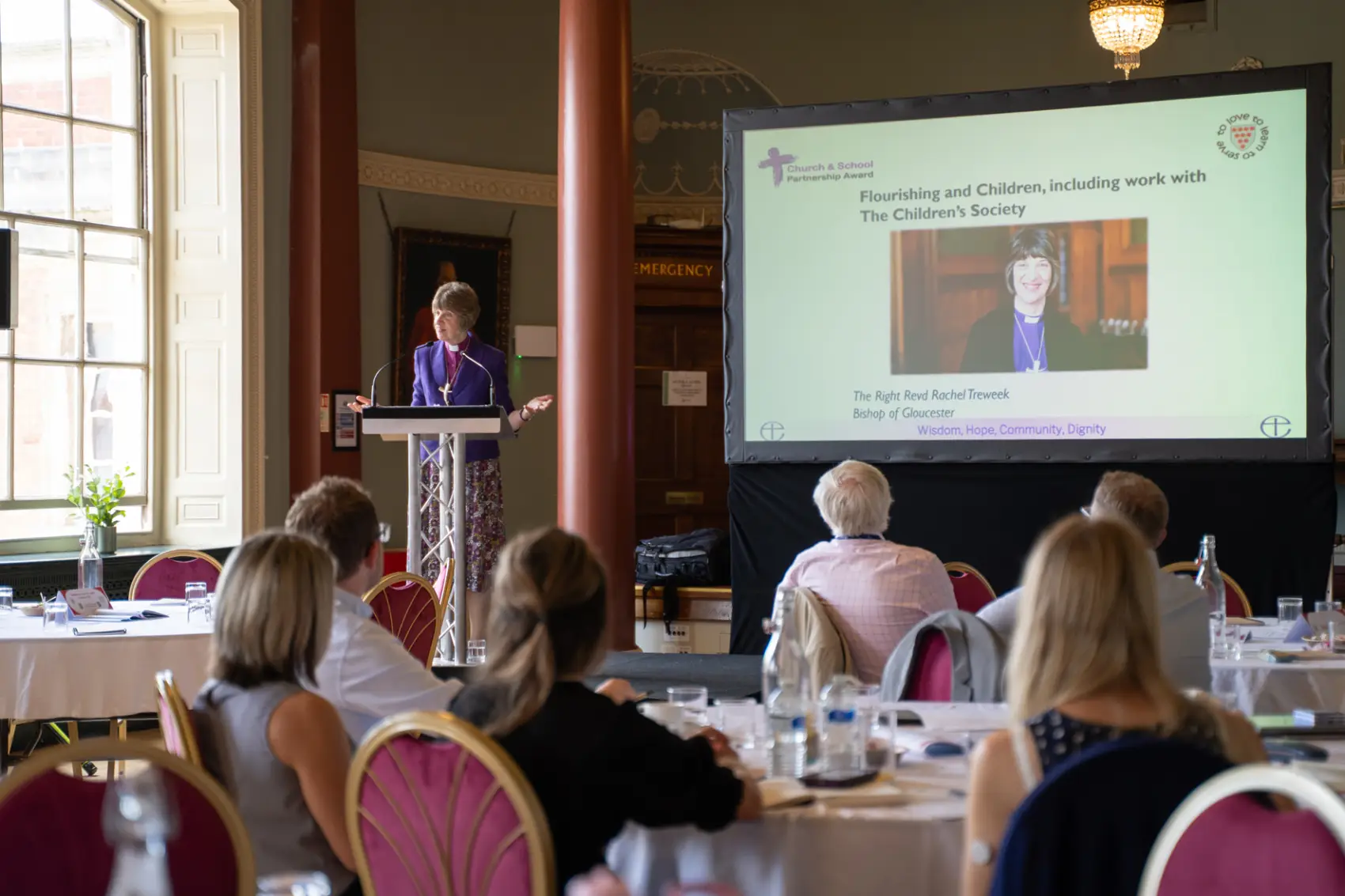 Bishop Rachel standing at a podium in front of a big screen while people sit around tables in front of her