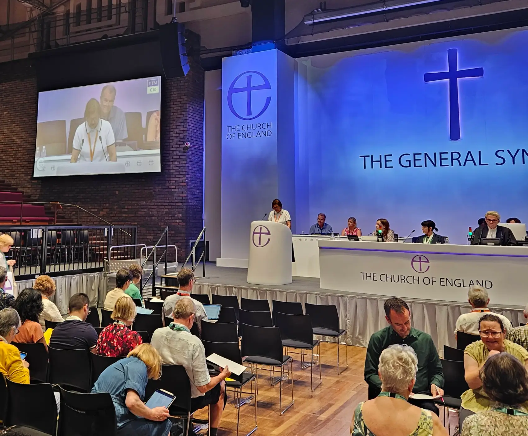 The inside of the General Synod chamber with people sat looking at a platform from which someone is speaking