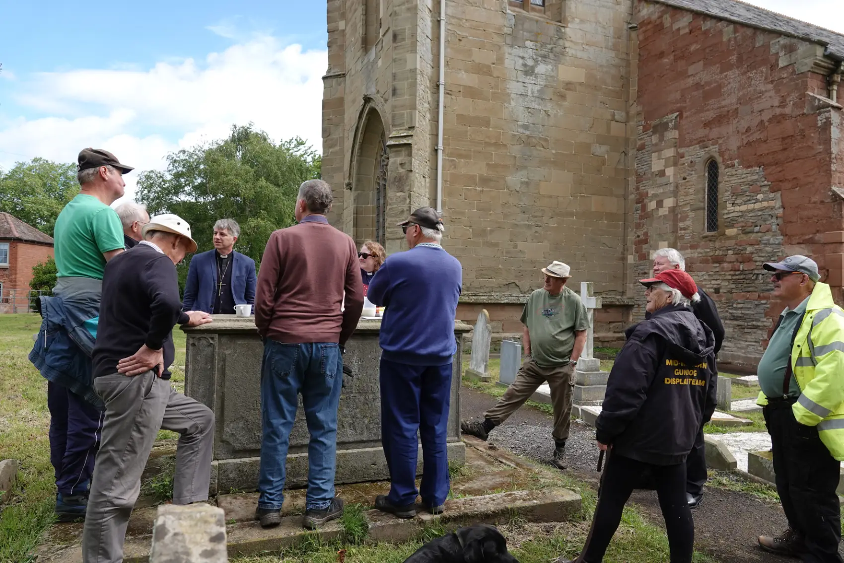 Bishop Hugh and members of the graveyard gang stand around an old grave chatting and drinking coffee