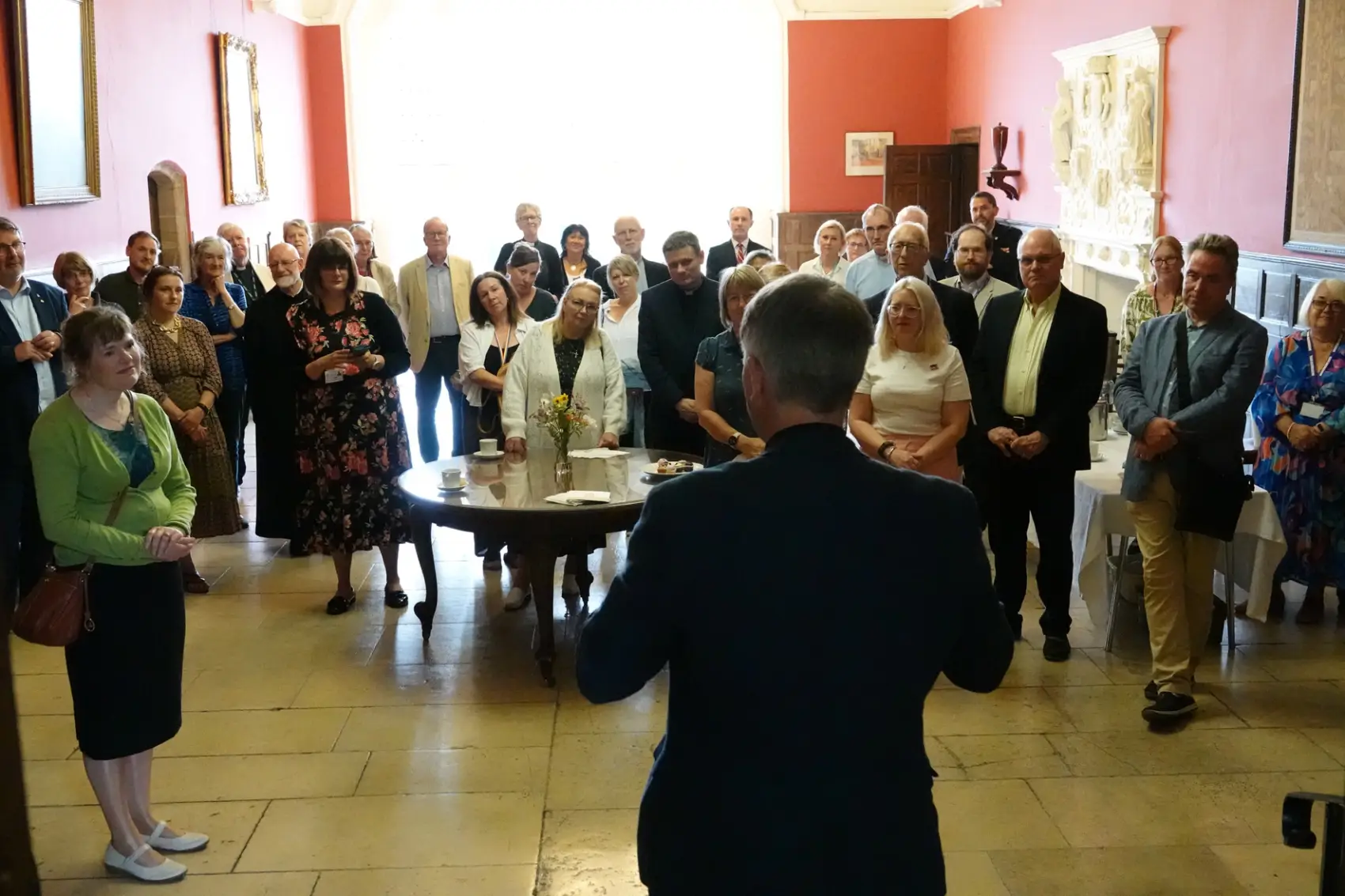 Looking towards a big window in the great hall, Bishop Hugh has his back to the camera as he talks to a group of gathered people