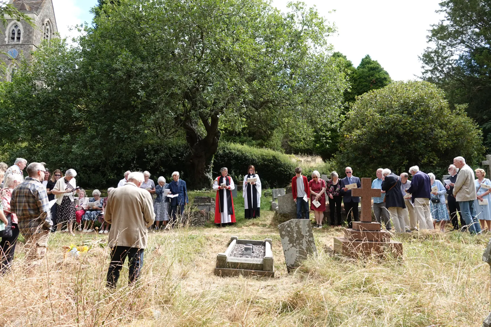 Bishop Hugh and Revd Rebecca stand in St James' Churchyard with a group of people either side of them. There are graves in the foreground